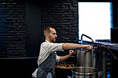 Young man in grey apron getting water from tap in big metal pan on restaurant kitchen