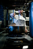 Young man in grey apron stirring big metal pan on restaurant kitchen