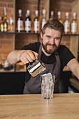 Cheerful bearded male barista in apron pouring ingredient from stainless pitcher into glass with ice cubes in coffee house