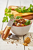 Ceramic bowl with salad of assorted nuts and vegetables with greens placed on white wooden bench with knife and fork