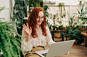 Positiv eingestellte rothaarige Mitarbeiterin mit Brille sitzt zwischen Zimmerpflanzen und winkt der Netbook-Kamera während einer Videokonferenz zu