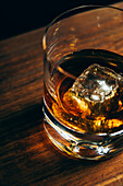 From above glass cup with cold whiskey and cube of ice placed on lumber table in dark room