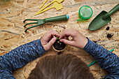 High angle of crop anonymous kid planting seedling in cardboard cup with ground at table with gardening shovel