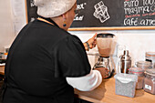 Woman in uniform and hat using grinder to prepare cocoa powder for hot drink during work in cafe