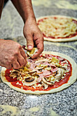 Nicht erkennbarer männlicher Koch, der bei der Arbeit in einer Restaurantküche geriebenen Käse auf einen Pizzaboden mit Tomatensauce streut