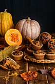 Composition made of appetizing muffins with chocolate chips placed on wooden table among scattered autumn leaves and pumpkins on dark background