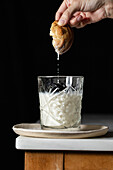 Crop anonymous person dipping fresh sweet cookie into glass of milk placed on corner of table against black background in kitchen