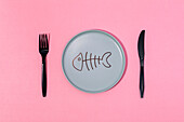 From above of gray ceramic plate with painted fish skeleton served on pink surface with black fork and knife