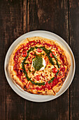 Von oben köstliche Pizza mit Kirschtomaten und Parmesan, garniert mit Mozzarella-Käsekugel und Pestosauce und serviert auf Teller auf Holztisch in Pizzeria