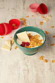 From above bowl of crunchy cornflakes and yogurt garnished with fresh raspberries and white chocolate placed on wooden table near red cupcake cases during breakfast