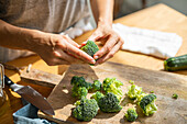 Crop anonymous cook preparing fresh green broccoli on wooden cutting board with knife while cooking in kitchen with bright sunlight
