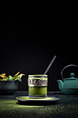 Still life composition with traditional oriental matcha tea served in glass cup with metal ornamental decor on table with ceramic bowls and fresh green leaves against black background