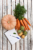 Draufsicht auf eine Ernte verschiedener frischer Gemüsesorten und einen geöffneten Notizblock mit Stift auf einem Holztisch