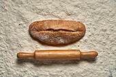 Top view full frame loaf of freshly baked bread with wooden rolling pin placed on white flour in light room