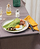 Hausgemachtes Frühstück aus Spinatpfannkuchen mit Speck, Ei und Pilzen, serviert auf einem weißen Teller mit einem Salz- und Ölstreuer auf einer karierten Tischdecke