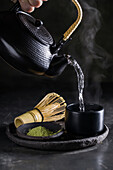 Crop anonymous person pouring hot water from kettle while preparing for tea ceremony with matcha powder on plate near chasen
