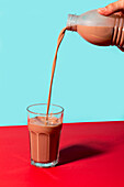 Unrecognizable person pouring sweet chocolate milk from plastic bottle into glass against two color background