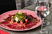 From above tomato salad with burrata and slices of prosciutto garnished with black olives and pesto sauce and served on plate on wooden table