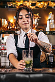 Young brunette in uniform mixing cold cocktail in glass cup while standing behind counter and working in bar