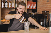 Serious bearded male barista in apron pouring ingredient from stainless pitcher into glass with ice cubes in coffee house