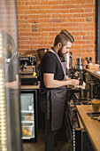 Seitenansicht eines bärtigen männlichen Barista, der einen Krug mit einer Serviette säubert, während er am Tresen neben der Kaffeemaschine steht und in einem Café arbeitet