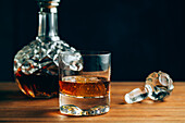 Glass of cold whiskey with ice placed on wooden table near decanter on black background