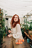 Charming young female with long wavy red hair standing with reusable string bag of tangerines in room with various houseplants