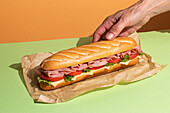 Cropped unrecognizable person hand holding appetizing sandwich with ham and fresh lettuce served on baking paper on colorful table background