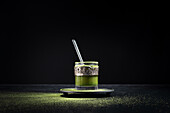 Gesunder grüner Matcha-Kräutertee, serviert in einer Glastasse mit Metallverzierung auf einer Untertasse, bestreut mit Pulver auf einem schwarzen Tisch