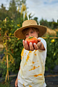 Blurred boy reaching out hand to camera with fresh red tomato while standing in rural garden in countryside during harvest season