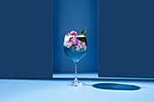 Transparentes Glas mit frischem Cocktail, Minzblättern und Blumen vor blauem Hintergrund auf dem Tisch