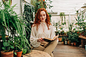 Concentrated red haired female sitting on chair with crossed legs and reading book among different potted plants in domestic garden