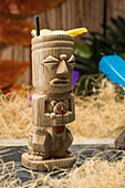 Traditional sculptural tiki cup of alcohol drink with straw placed on rug against wooden fence colorful leaves and dry grass