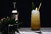 Yellow cocktail in glass garnished with pineapple piece and green leaves with paper straw near bar spoon