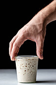 Hand of crop anonymous person demonstrating white round glass cup with sourdough starter and covered with glass lid placed on table against black background
