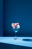 Transparentes Glas mit frischem Cocktail, Minzblättern und Blumen vor blauem Hintergrund auf dem Tisch