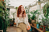 Charming young female with long wavy red hair standing with reusable string bag of tangerines in room with various houseplants