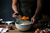 Cropped unrecognizable male cook using wooden spoon to mix pumpkin puree with eggs and flour in bowl while preparing pie on timber table near cutting board and towel