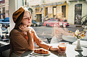 French female in beret sitting at table in cafe with aromatic glass of coffee and freshly baked croissant