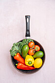 Top view of frying pan with raw ripe peppers and avocados near cherry tomatoes and greens on light background in kitchen