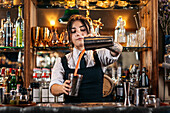 Female barkeeper pouring orange alcohol cocktail in metal shaker while preparing refreshing drink and standing at counter in bar