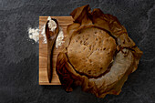 Komposition von oben mit frisch gebackenem, rustikalem, rundem Sauerteigbrot auf Pergamentpapier, das auf einem Holzbrett mit Löffel und Weizenmehl liegt