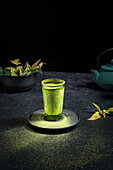 Still life composition with traditional oriental matcha tea served in glass cup with metal ornamental decor on table with ceramic bowls and fresh green leaves against black background