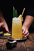 Crop anonymous barman garnishing alcohol cocktail with green leaves and pineapple piece on tray near shot glass at wooden table on black background