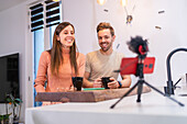 Positive couple of bloggers recording vlog on smartphone with microphone while sitting at kitchen counter with cup of coffee at home
