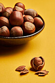 Fresh unpeeled hazelnuts placed in bowl near cracked nut on yellow background