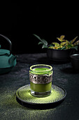 Still life composition with traditional oriental matcha tea served in glass cup with metal ornamental decor on table with ceramic bowls and fresh green leaves against black background