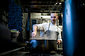 Junger Mann in grauer Schürze rührt in einer großen Metallpfanne in einer Restaurantküche