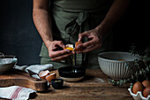 Unrecognizable guy in apron breaking raw egg over bowl while preparing pastry on lumber table near kitchenware