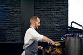 Junger Mann in grauer Schürze rührt in einer großen Metallpfanne in einer Restaurantküche